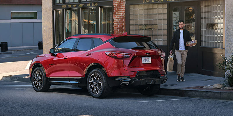 Red 2026 Chevrolet Blazer SUV parked on a city street outside a storefront, featuring sporty styling, black wheels, and a rear three-quarter view as a person walks out carrying groceries.